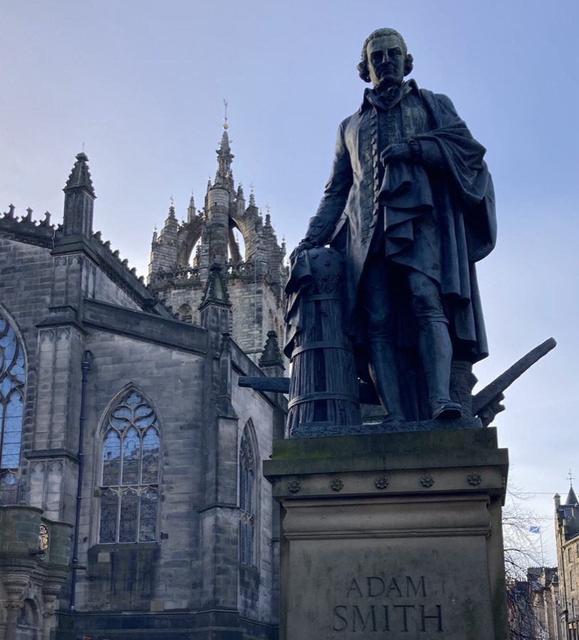 Bronze statue of Adam Smith on the Royal Mile in Edinburgh, standing in eighteenth-century dress with a hand on a beehive and plough, St Giles' Cathedral behind him under a blue sky.